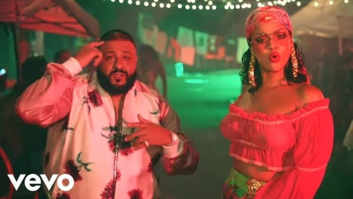 A man and a woman stand under string lights at night; the man gestures to his head while the woman, channeling Rihanna in "Wild Thoughts," wears sunglasses, a headscarf, and a red off-shoulder top.
