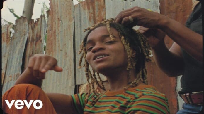 A person with blonde-tipped dreadlocks sits outdoors in front of a corrugated metal fence while another styles their hair. The Vevo logo and “Koffee Toast Official Video” branding are visible in the bottom left corner.