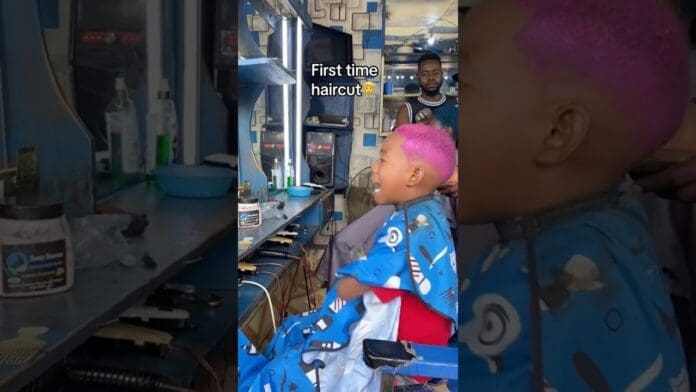 A young child with bright pink hair sits in a barber chair for a haircut, wearing a blue cape, while a barber stands in the background. Text reads “First time haircut.”—a 2024 remix challenge inspired by fresh Amapiano vibes.