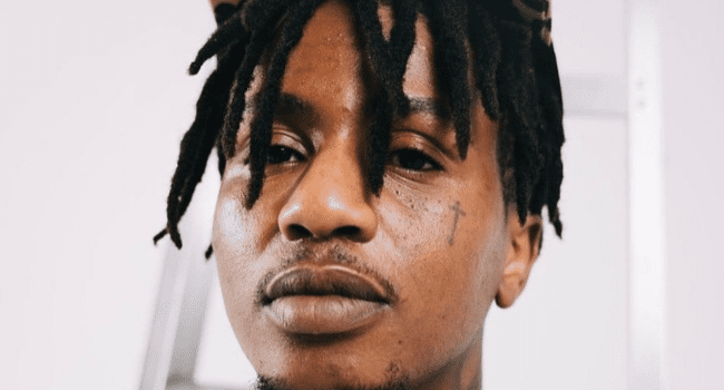 A person with short dreadlocks, a faint face tattoo, and a mustache looks directly at the camera against a plain background, reminiscent of Emtee after dozing off on stage.