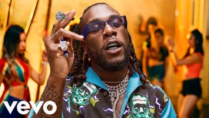 A man wearing sunglasses, jewelry, and a colorful shirt gestures with his hand as several people dance in the blurred background. The Vevo logo is visible in the corner, giving the scene a vibe reminiscent of a Chris Brown music video.