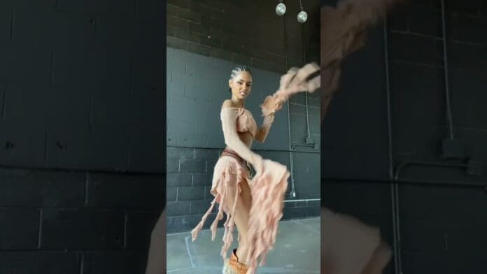 A woman in a peach, tattered dress poses in an industrial-style room with black walls and exposed pipes—giving major freshoutthegallery vibes perfect for an Album drops promo or YouTube visual.