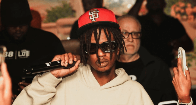 Person wearing sunglasses and a red SF cap holds a microphone and gestures with their hand during a live performance at an indoor event; people are visible in the background.