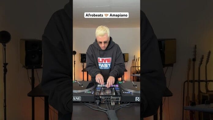 A person with blond hair wearing sunglasses and a "LIVE FAST" hoodie skillfully mixes Afrobeats on DJ equipment; guitars are visible in the background, highlighting a smooth transition between genres.
