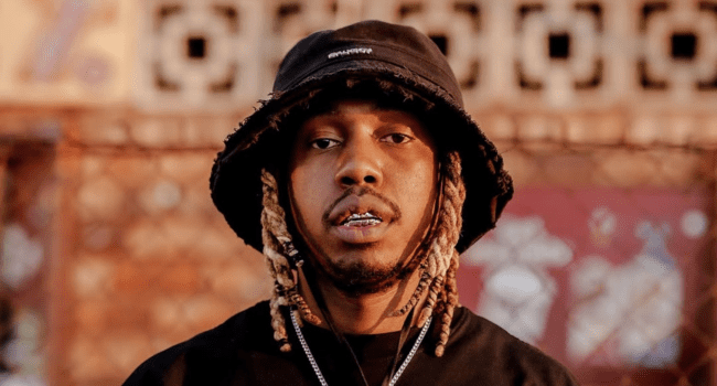 A man wearing a black bucket hat, black shirt, and necklace stands outdoors, looking at the camera. With light brown dreadlocks and silver grillz, he showcases the evolving style of SA Hip-Hop as it approaches new historical milestones in 2025.