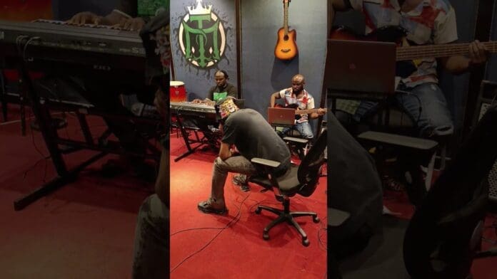 Four people are in a music studio; one plays keyboard, one plays guitar, another does a freestyle on a laptop, and one sits facing away. Musical instruments hang on the wall, capturing an I Hail Una vibe inspired by 2Baba.
