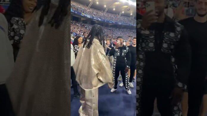 A person in a shiny, oversized outfit stands facing a group of people in a crowded stadium during the Champions League final, with spectators in the background, capturing an iconic afrobeat-inspired moment.