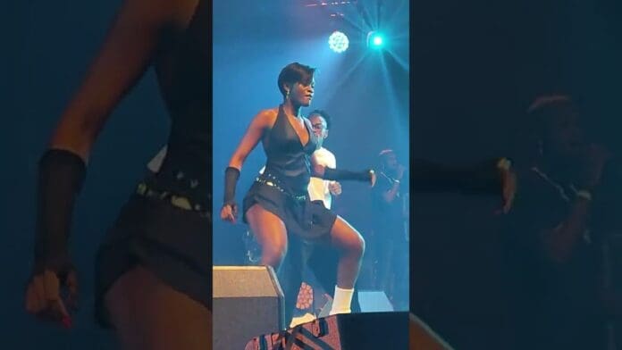 Two performers dance energetically on stage under colorful lighting at a Fally Ipupa concert, with musicians and equipment visible in the background.