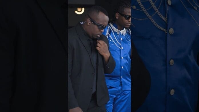 Two men walk side by side; one, Munyakazi, sports a black suit and sunglasses, while the other stands out in a bright blue suit with silver chains and sunglasses.