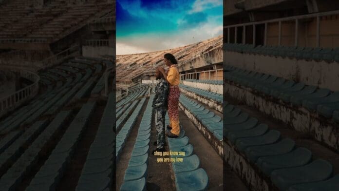 Two people stand closely together in an empty stadium with blue seats, facing each other under a partly cloudy sky. Yellow text at the bottom reads, "shey you know say you are my love," capturing a lucid, Afrobeats-inspired moment.