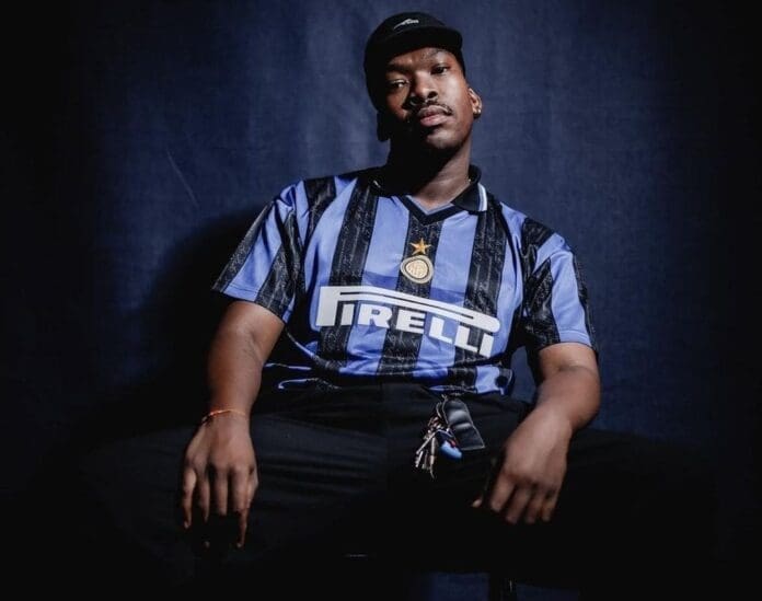 A person sits against a dark background wearing a black cap and a blue and black striped Inter Milan football jersey with a Pirelli sponsor logo, reminiscent of the laid-back style seen among Amapiano artists like Thuly Thulz.