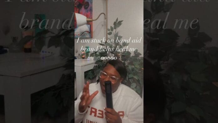 A person in a white sweater holds a microphone and sings the catchy “I am stuck on band aid brand helps heal me ooo oo,” next to a plant indoors, blending the vibe of latest song music 2025.