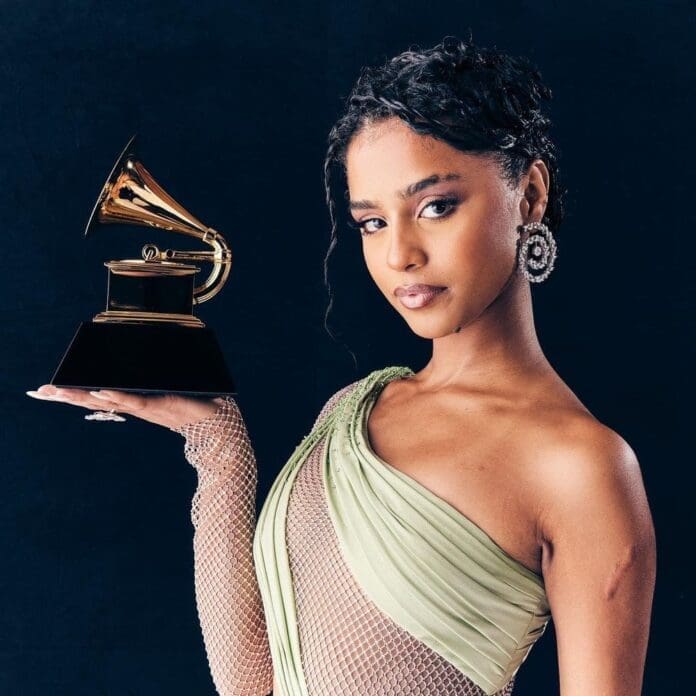 Tyla, in a light green dress, holds a Grammy Award trophy against a dark background, looking at the camera.