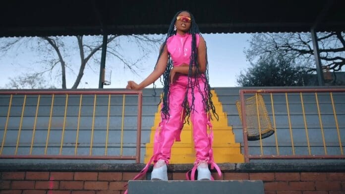 Person standing on stairs in an outdoor stadium, channeling Zuchu vibes in a bright pink outfit, platform shoes, and sunglasses, with long braids and trees in the background—perfect for an Official Music Video look.