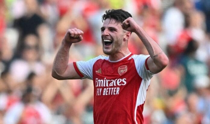 Declan Rice, in an Arsenal jersey, celebrates with both fists raised and a wide smile, on a field with a blurred crowd in the background.