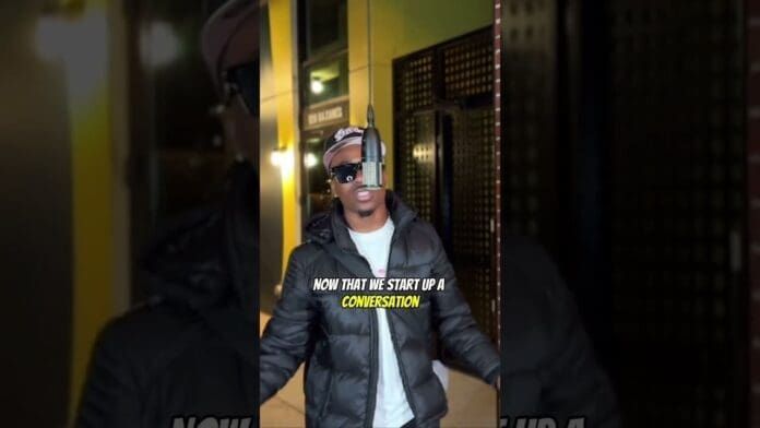 A person wearing sunglasses and a puffer jacket speaks into a microphone at an open mic outdoors at night. Text on the image reads, "NOW THAT WE START UP A CONVERSATION.