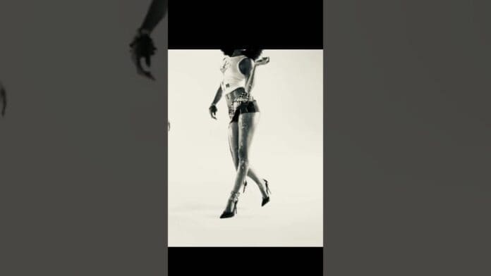 Black and white image of a woman in a tank top, shorts, and high heels, posing with arms raised and head turned away; her face is not visible, capturing the bold energy of popmusic.