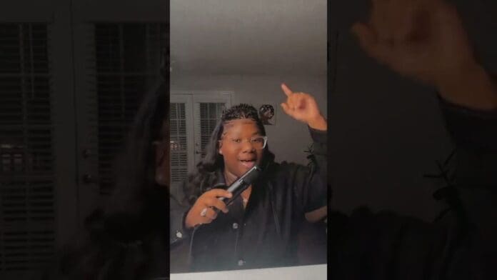 A person holding a hair straightener and raising one hand while standing indoors in front of a window, as afrobeats latest songs play softly in the background.