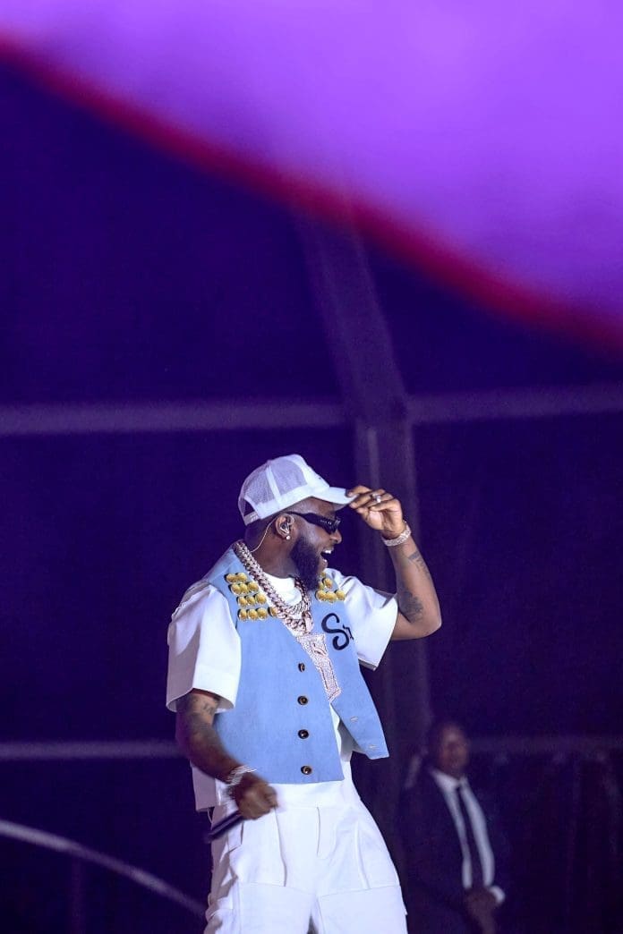 A man wearing a light blue vest, white shirt, white cap, and gold chains performs on stage at the Davido Concert in Abuja, with a security guard visible in the background.