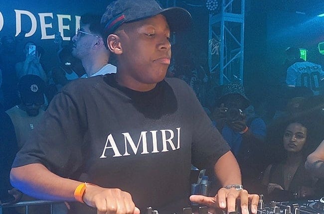 A DJ wearing a black Amiri t-shirt and cap performs an amapiano set at a club, with a crowd and blue lighting in the background.