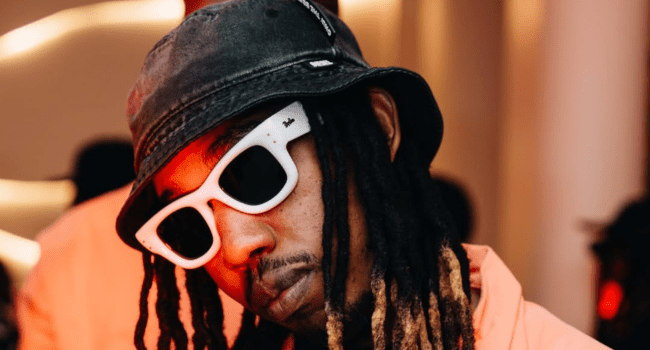 Person wearing a black bucket hat, white sunglasses, and a peach shirt, with long dreadlocks—channeling Best Hip Hop Album vibes like 25K at the SAMAs—looks confidently at the camera in an indoor setting.