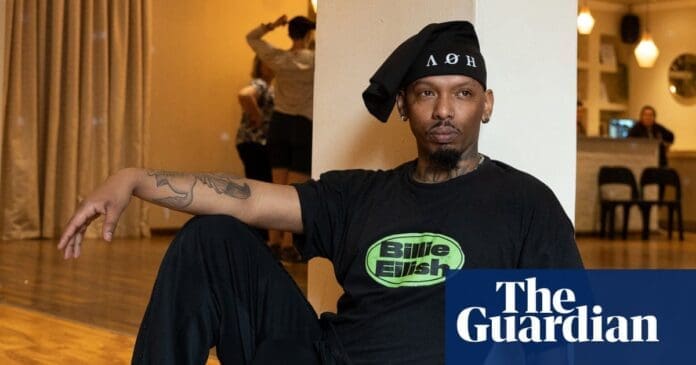 A man sits on a wooden floor in a dance studio, wearing a Billie Eilish t-shirt and a black headscarf; two people dance in the background. The Guardian logo is visible, highlighting the global reach of this South African choreographer.