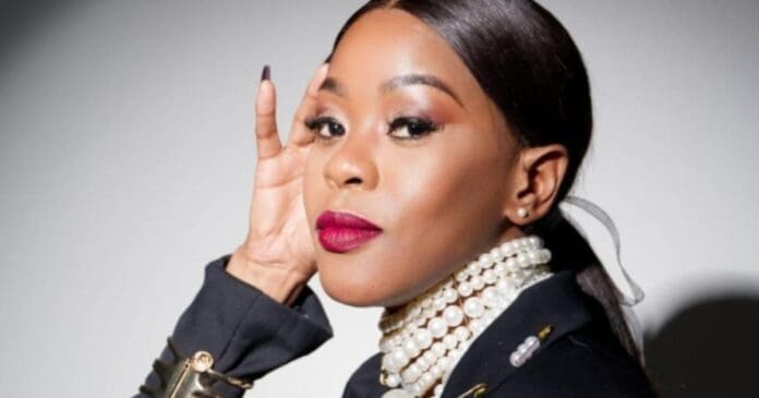 A woman with straight dark hair in a low ponytail, wearing a black outfit, layered pearl necklace, and bold red lipstick, poses with her hand lifted near her face in true Naima Kay style, echoing the elegance of maskandi influences.