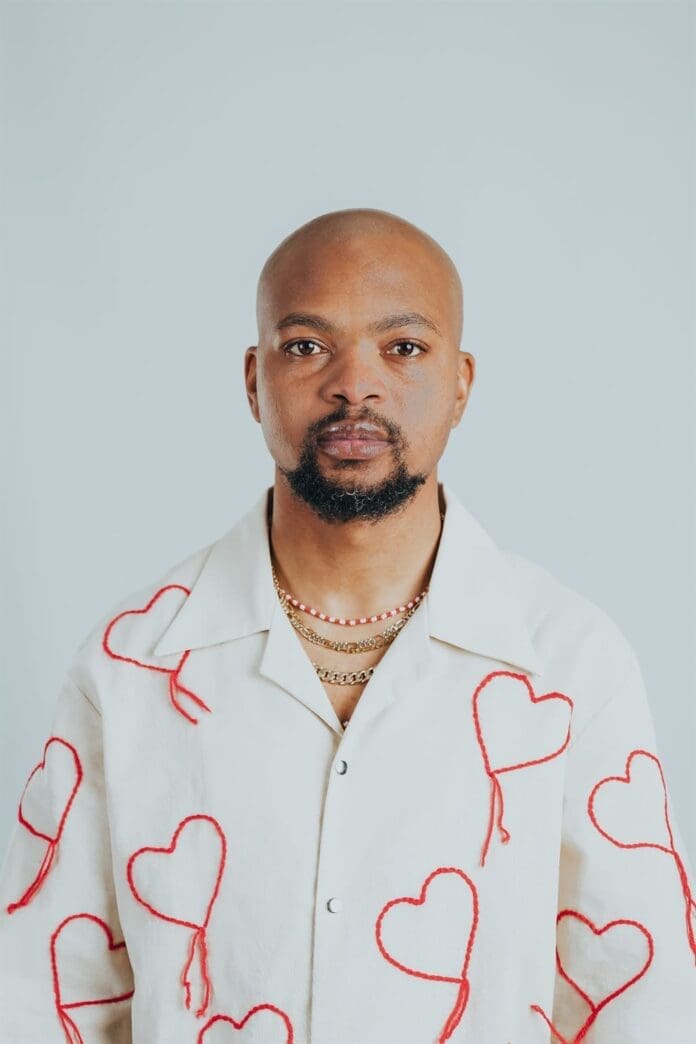 Amapiano star with a shaved head and beard wears a white shirt with red heart designs and layered necklaces, standing against a plain light background.
