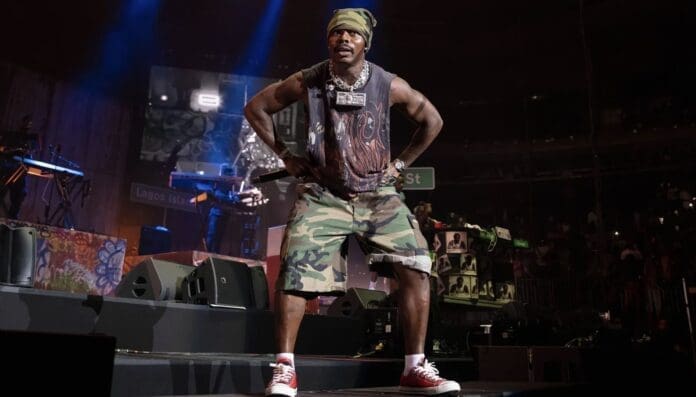 A man in camo shorts, a green headwrap, and red sneakers stands on stage holding a microphone, channeling vibes that would fit perfectly on the Spotify African Heat Playlist. Stage lights and musical equipment glow in the background.