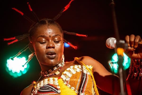 A woman with beaded hair, facial paint, and a shell necklace sings on stage, holding a microphone with colorful lights in the background—reminiscent of the vibrant energy of One Corner.