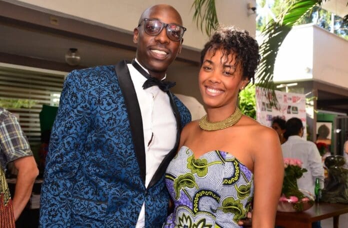 A man in a blue patterned suit and bow tie stands next to a woman in a patterned strapless dress, both smiling at an indoor event celebrating Bien and his chart-topping songs.