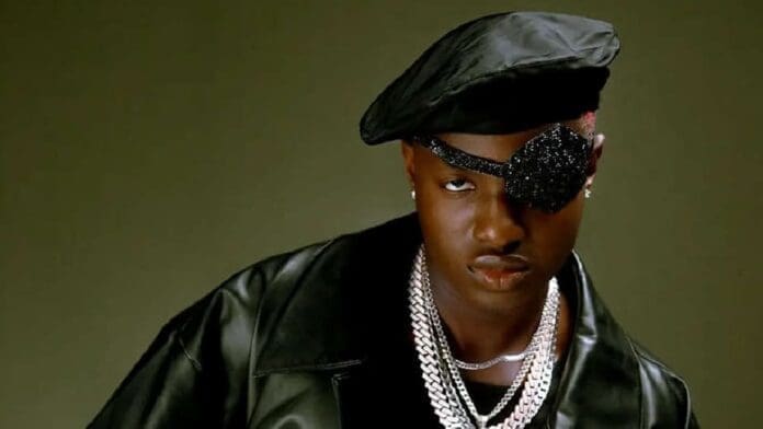 A person wearing a black leather jacket, a black beret, an embellished eye patch, and layered silver chains poses confidently against a plain background, channeling Ruger’s unique Afropop style.