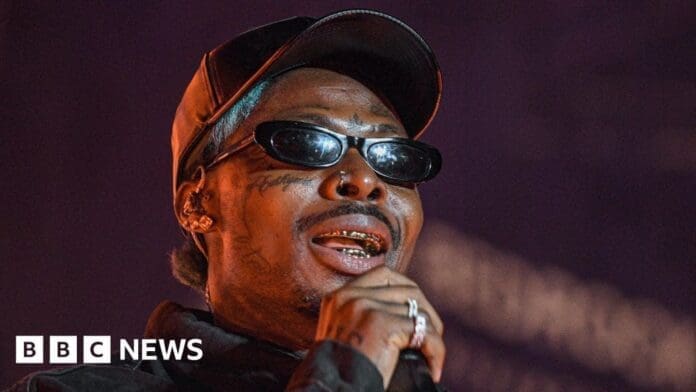 A person wearing dark sunglasses, a cap, and gold dental grills—resembling a Nigerian Afrobeats star—performs on stage under purple lighting. The BBC News logo is visible in the corner.