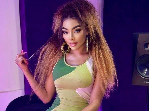 A woman with long, curly brown hair wearing a green and beige dress sits indoors, holding a strand of her hair, with a speaker in the background—her vibe echoing Reggae fusion and Afro-pop influences.