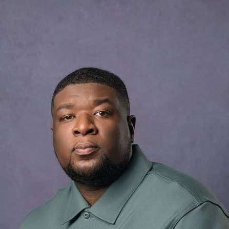 A man with short black hair and a trimmed beard wearing a green collared shirt poses in front of a plain grey background, capturing the contemporary vibe of Ghanaian Music.