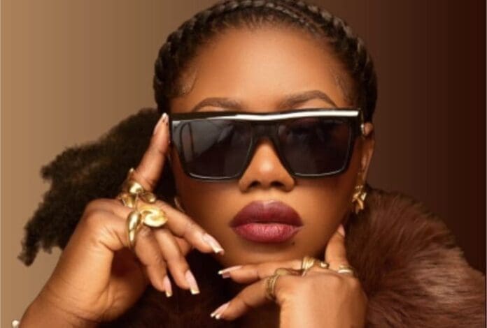A woman with braided hair wears large black sunglasses, gold rings, and hoop earrings, striking an OMOG3-inspired pose with her hands near her face against a brown background.