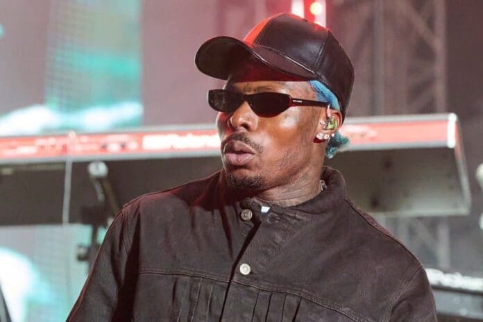 A man wearing a black cap, sunglasses, and a dark jacket performs on stage, with a keyboard visible in the background at an Asake concert featured by Billboard.