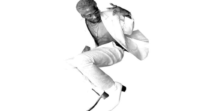 A person with bleached hair wearing a white suit, sunglasses, and jewelry poses mid-air against a white background with their jacket open, embodying the bold energy of afrobeats.