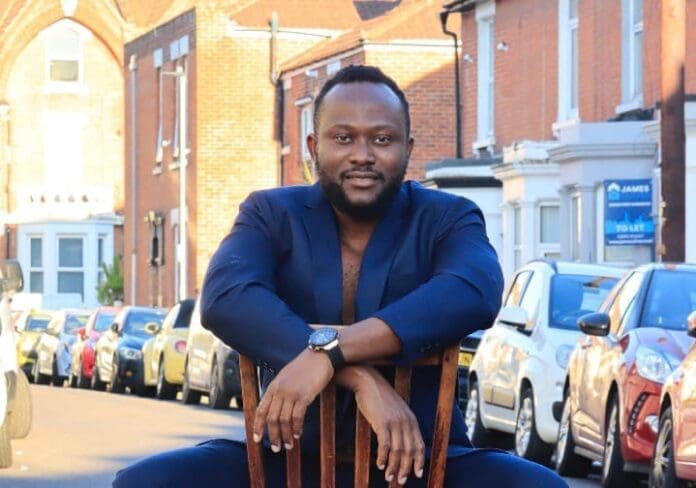 A man in a dark blue suit sits on a wooden chair in the middle of a residential street, where street music echoes between parked cars and brick houses, reminiscent of scenes covered by The Guardian Nigeria.