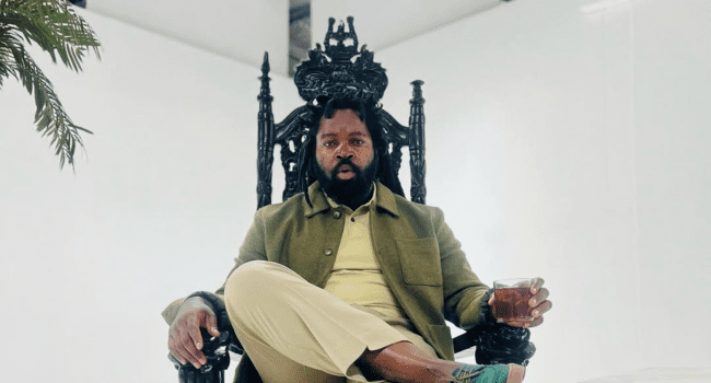 A man with a beard sits on an ornate black throne, holding a drink in one hand, wearing a green jacket and beige pants, in a white room with a plant on the left—evoking the commanding presence of Big Zulu after the Bergville Soccer Tournament.