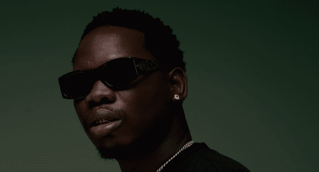 A man wearing dark sunglasses, a black shirt, a silver chain, and an earring poses against a plain dark green background, channeling the bold vibe of a Clout Cassette music video.