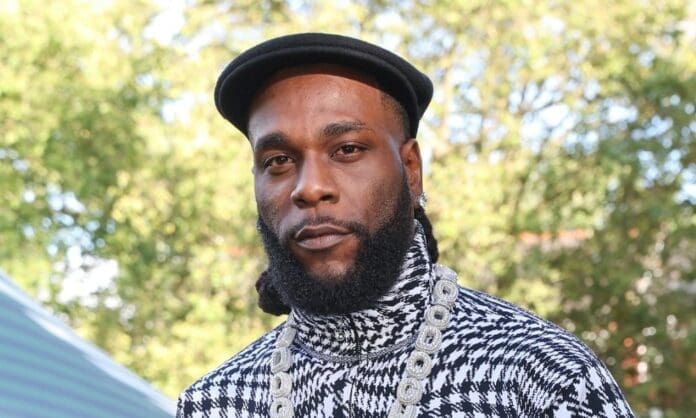 A man wearing a black beret, patterned turtleneck, and large chain necklace stands outdoors with trees in the background, channeling Burna Boy’s iconic style—one of Africa’s biggest stars behind the highest-grossing music tour.