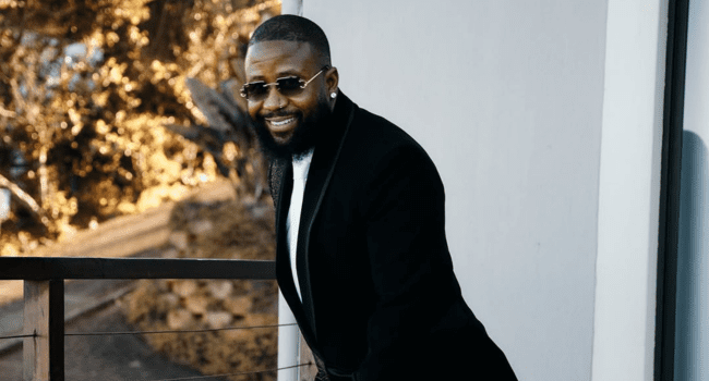 A man wearing a black suit and sunglasses stands on a balcony, smiling as if celebrating Cassper’s plan to Fill Up Toyota Stadium, with trees visible in the background.