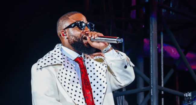 A man wearing sunglasses, a white coat with black polka dots, and a red tie performs on stage with a microphone at the Cassper Fill Up event in Toyota Stadium.