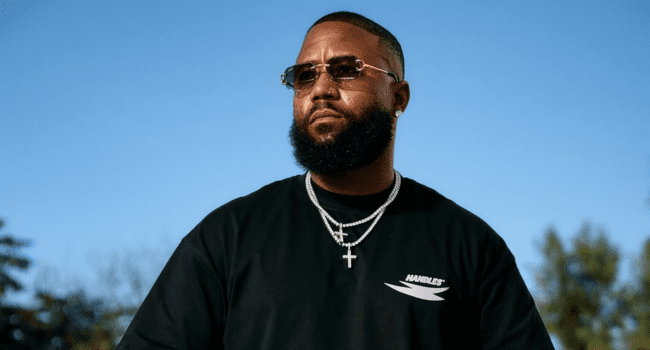 A man with a beard and sunglasses, channeling Cassper’s style, wears a black t-shirt and layered silver necklaces while standing outdoors with a blue sky in the background.