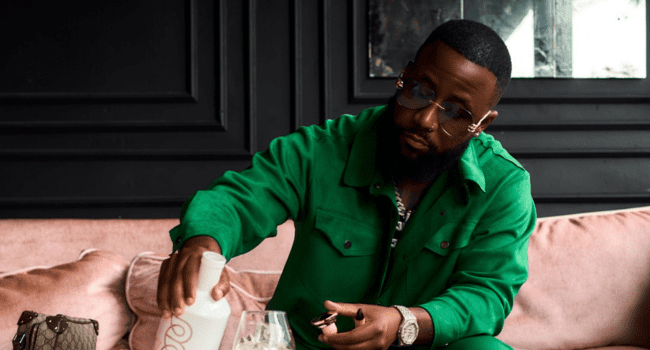 A man in a green outfit sits on a pink couch, pouring a drink into a glass while holding a cigar, celebrating his 35th Birthday with gratitude.