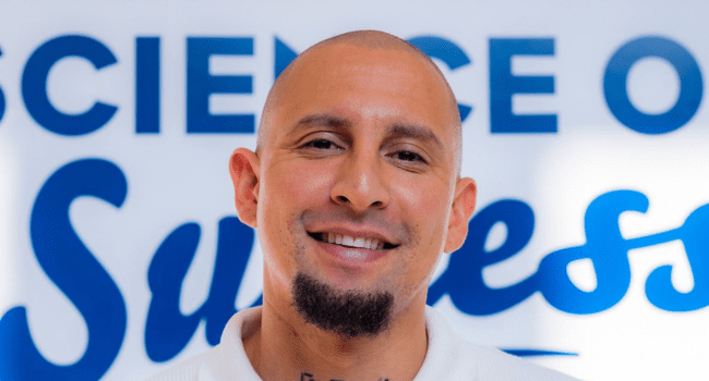 A man with a shaved head and goatee, reminiscent of DJ Warras, smiles in front of a blurred sign that reads "SCIENCE OF SUCCESS," capturing the spirit of SA Hip Hop achievement.
