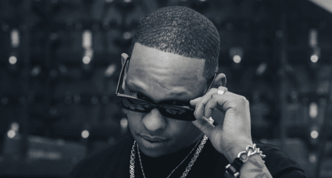 A person wearing sunglasses and jewelry, reminiscent of Da LES’s style, looks over the top of their sunglasses in a dimly lit setting.