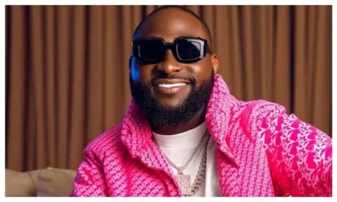 A man with a trimmed beard wears black sunglasses, a white shirt, and a pink patterned jacket. He is smiling and sitting indoors in front of brown curtains, resembling the stylish vibe often seen in Daily Post Nigeria features on artists like Davido.