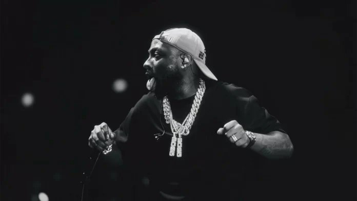 A man wearing a backwards cap and multiple chains performs on stage, sticking out his tongue and holding a microphone, with a dark background—capturing the wild energy of Detty December and the vibes of 2025 anthems.